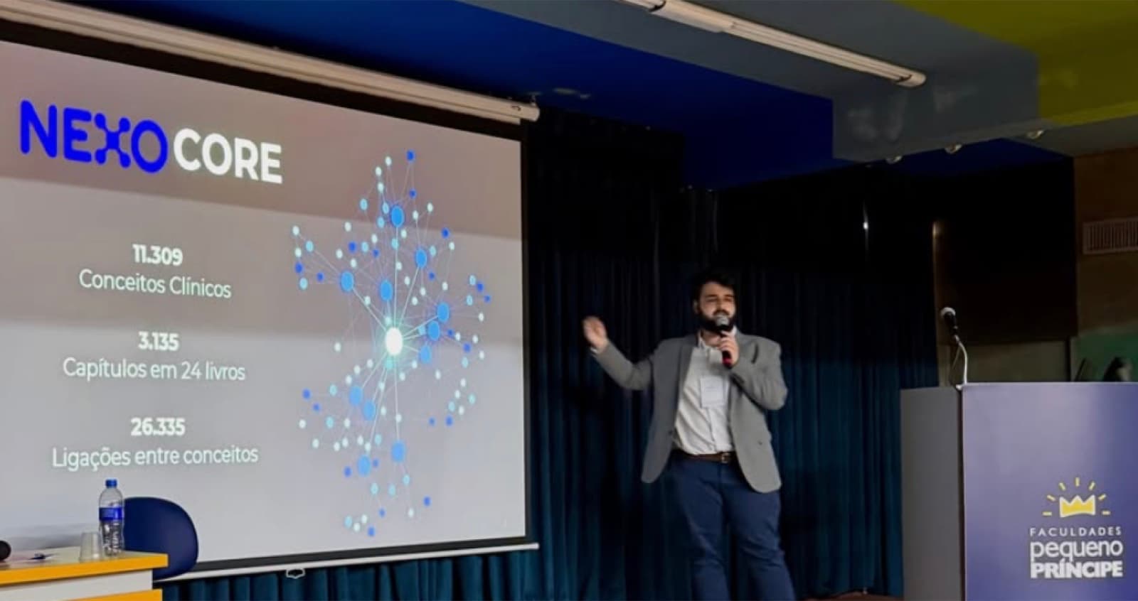 Thiago Moraes ao microfone diante de um telão com o slide do NEXO Core e um grafo de conceitos clínicos, no auditório das Faculdades Pequeno Príncipe.
