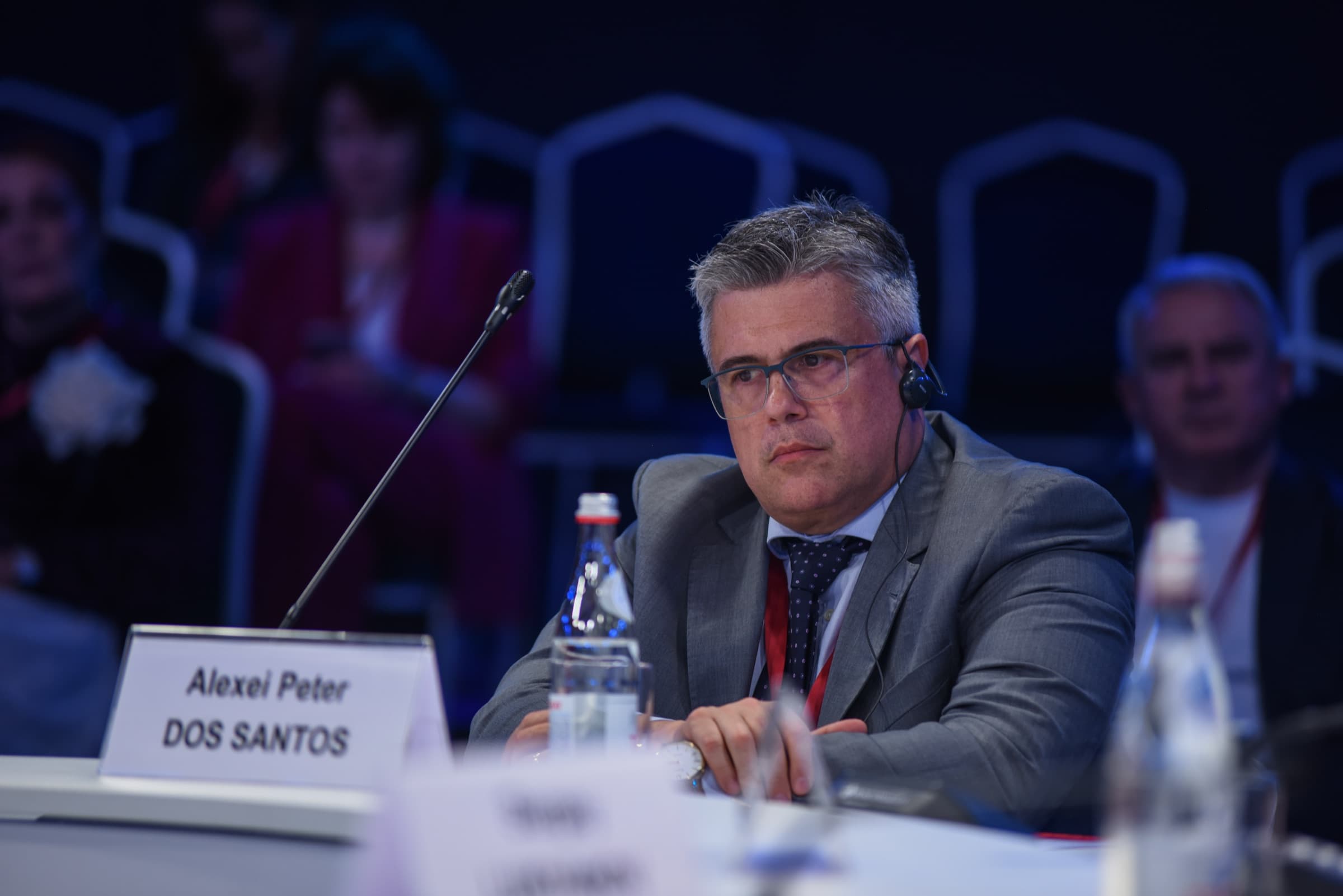 Dr. Alexei Peter sentado à mesa da sessão, com fone de tradução simultânea e sua placa de identificação à frente.